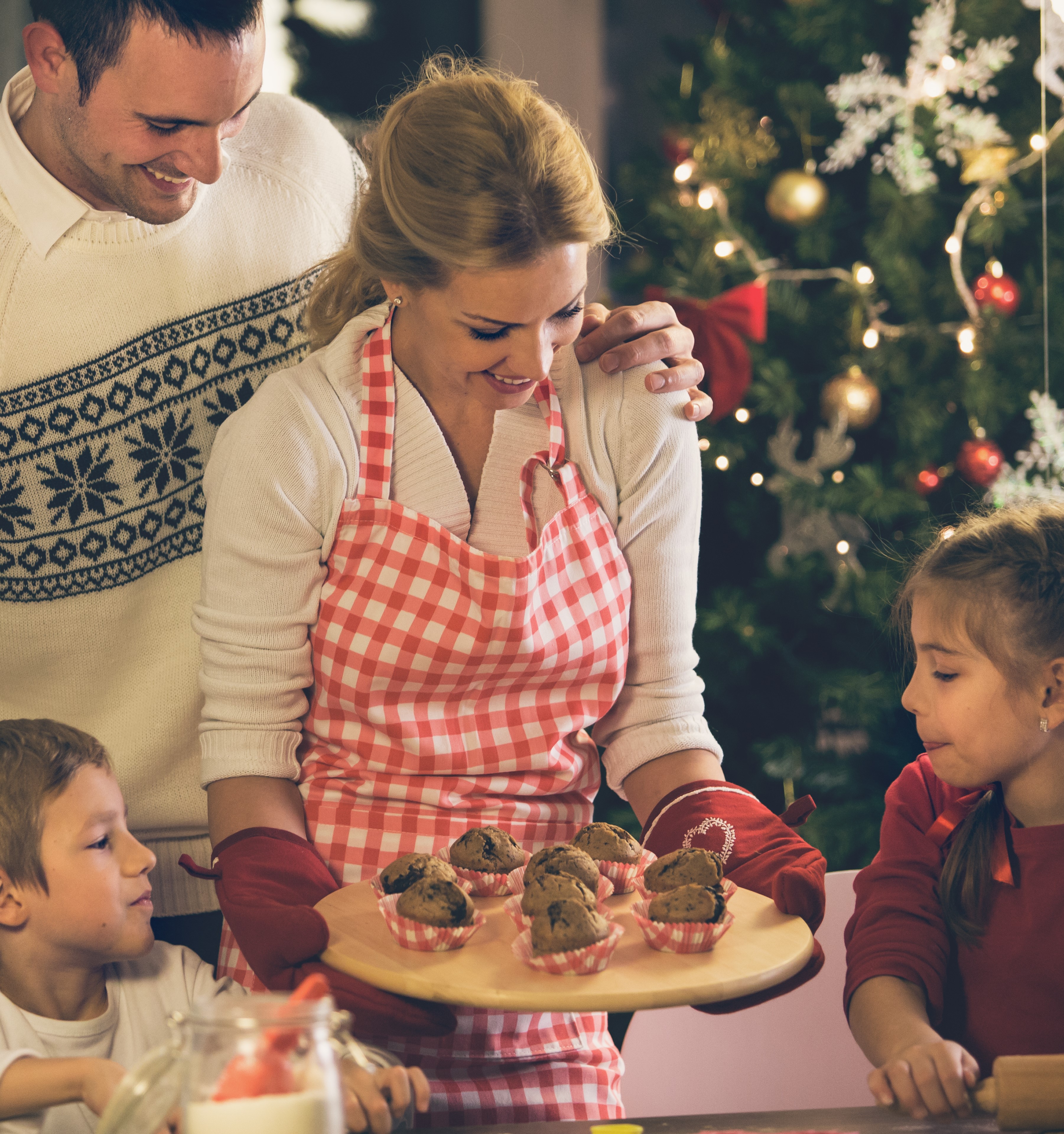 A man embraces his wife as she holds a tray of healthy holiday muffins for their children in front of a decorated Christmas tree.
