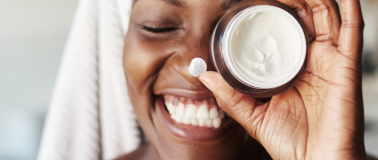 A woman hold up a small jar of moisturizer to her left eye while smiling
