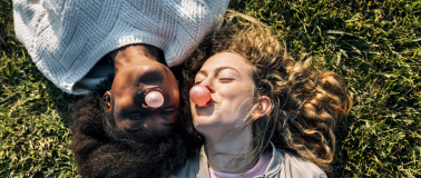A close-up of two women laying in a grassy field blowing bubble gum bubbles